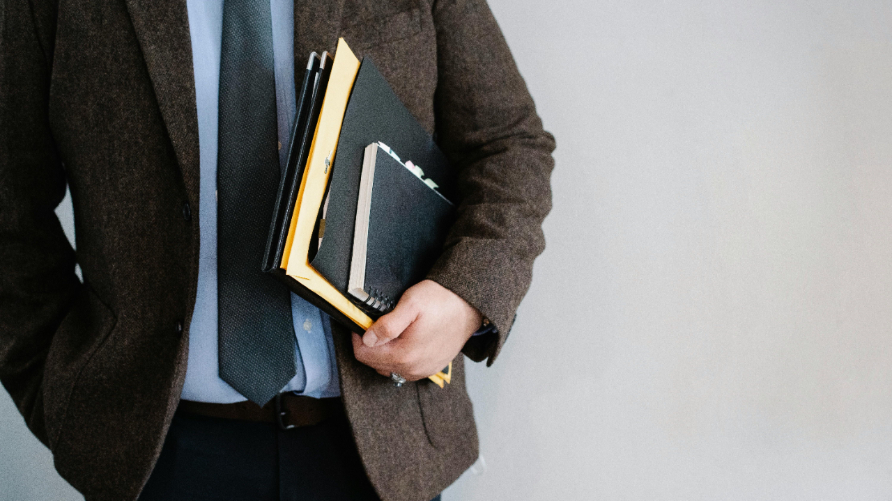 A person in a brown blazer holds a stack of notebooks and folders against a plain gray background.